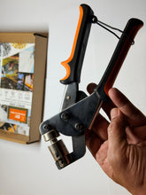 Hand holding a black and orange tool against a white wall with a box and images in the background.